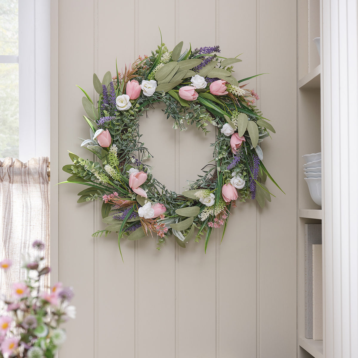 Frühlingskranz mit rosa Tulpen, Lavendel und weißen Rosen an einer hellen Wand neben einem Fenster