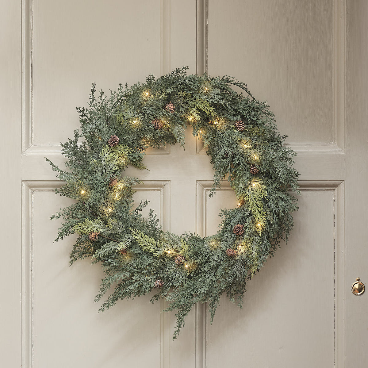 Nahaufnahme des LED-Weihnachtskranzes mit Tannenzapfen und warmweißen LEDs an einer hellen Tür