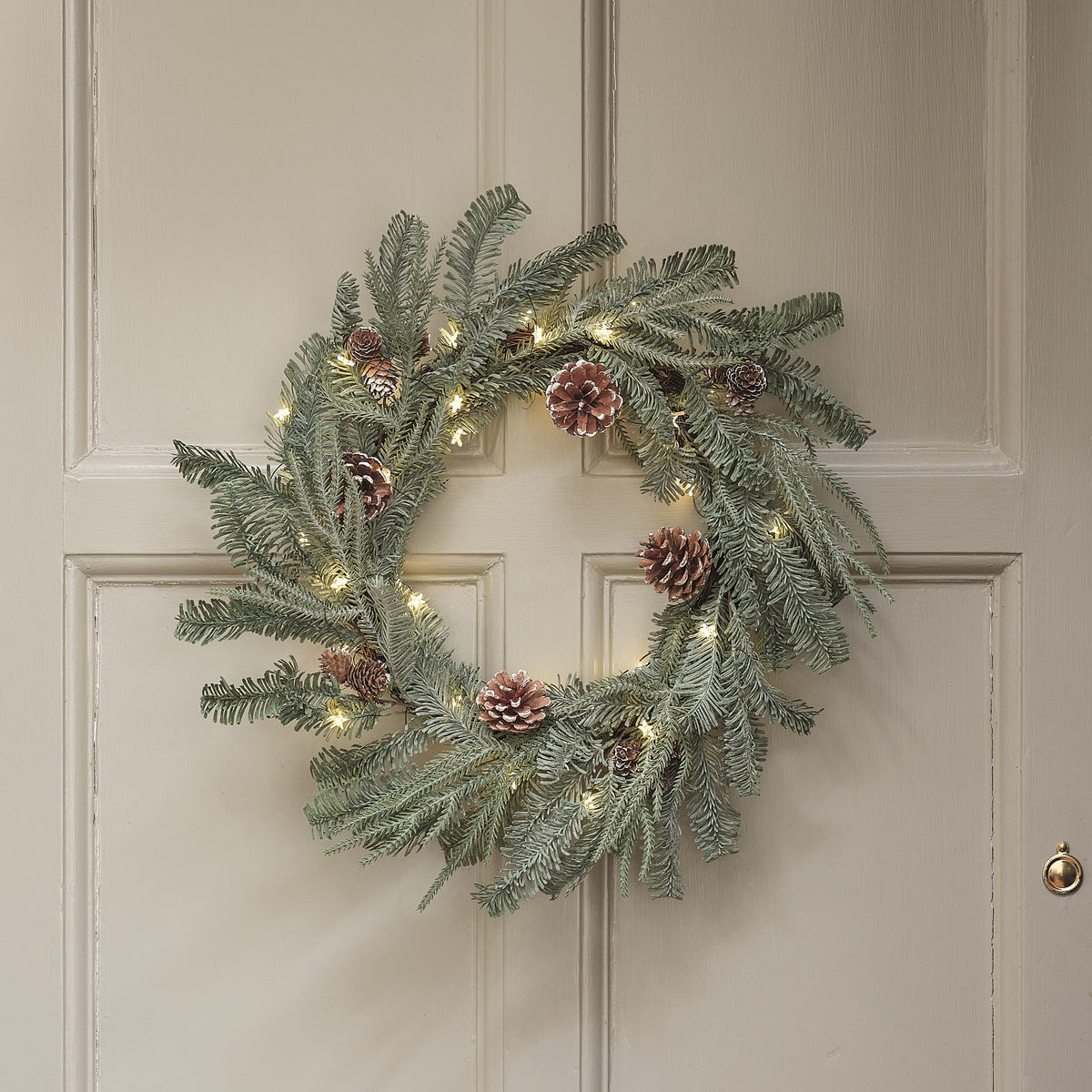 Beleuchteter Weihnachtskranz mit Zapfen an weißer Tür hängend