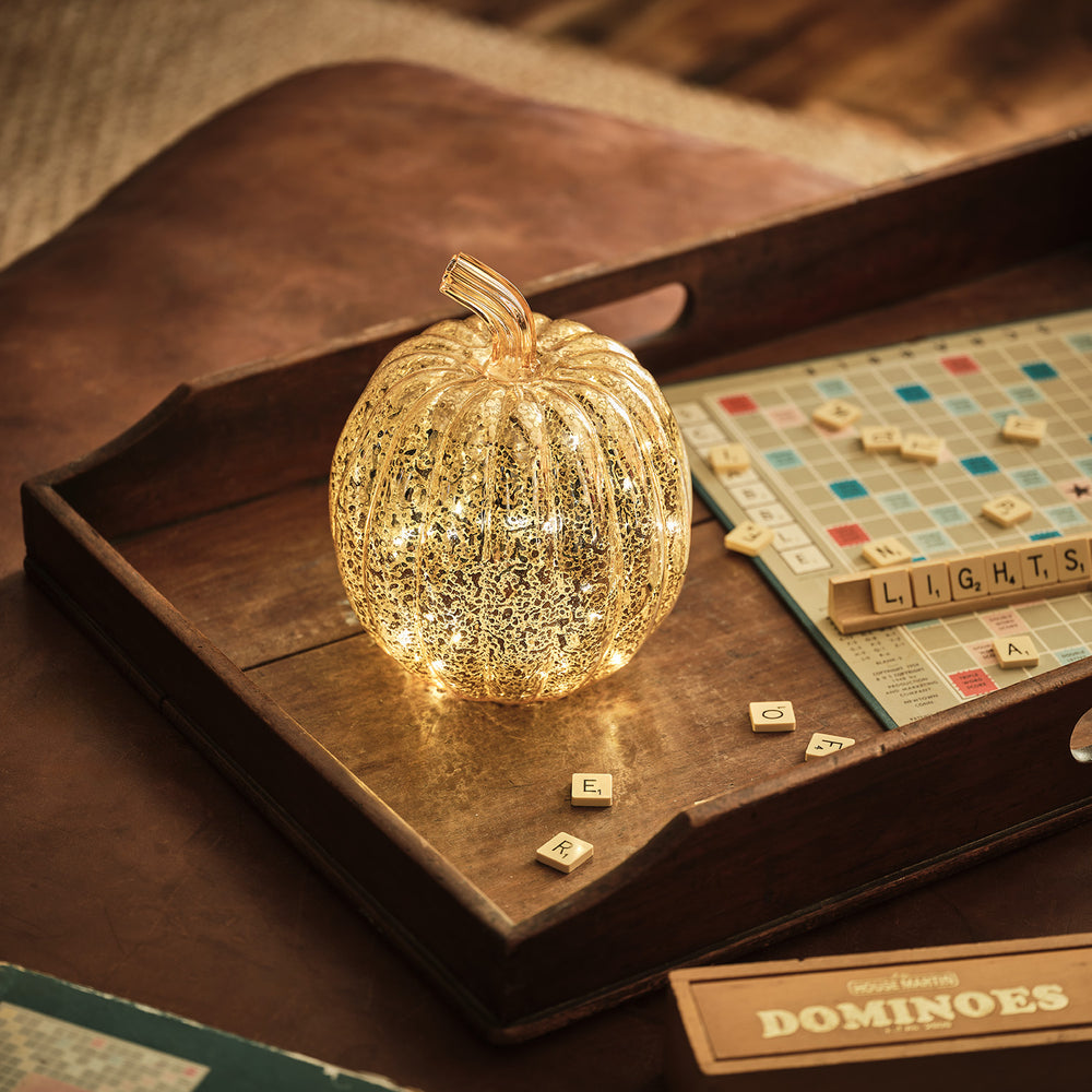 Leuchtender antikgoldener Glas-Kürbis auf Holztablett neben Scrabble-Spiel