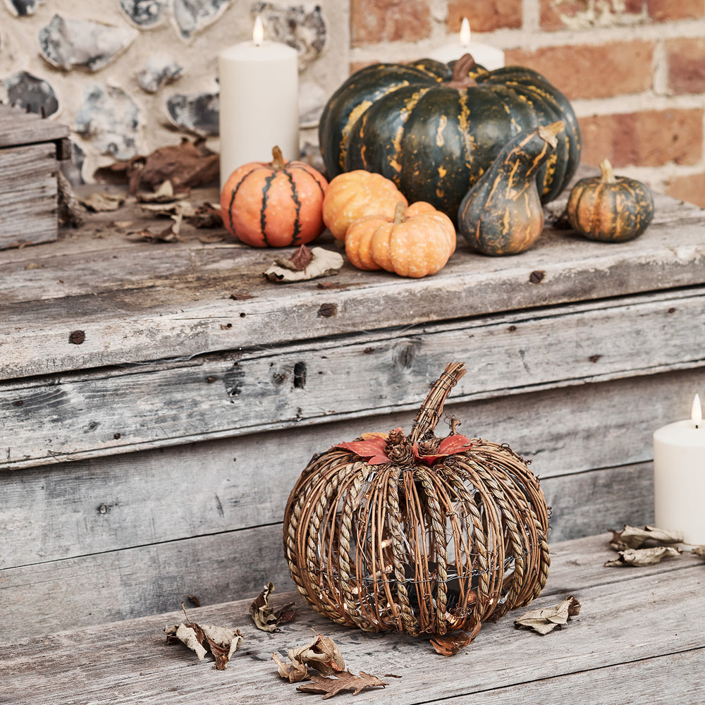 Rattan-Kürbis mit Herbstdeko und weißen Kerzen auf verwittertem Holztisch vor Steinwand