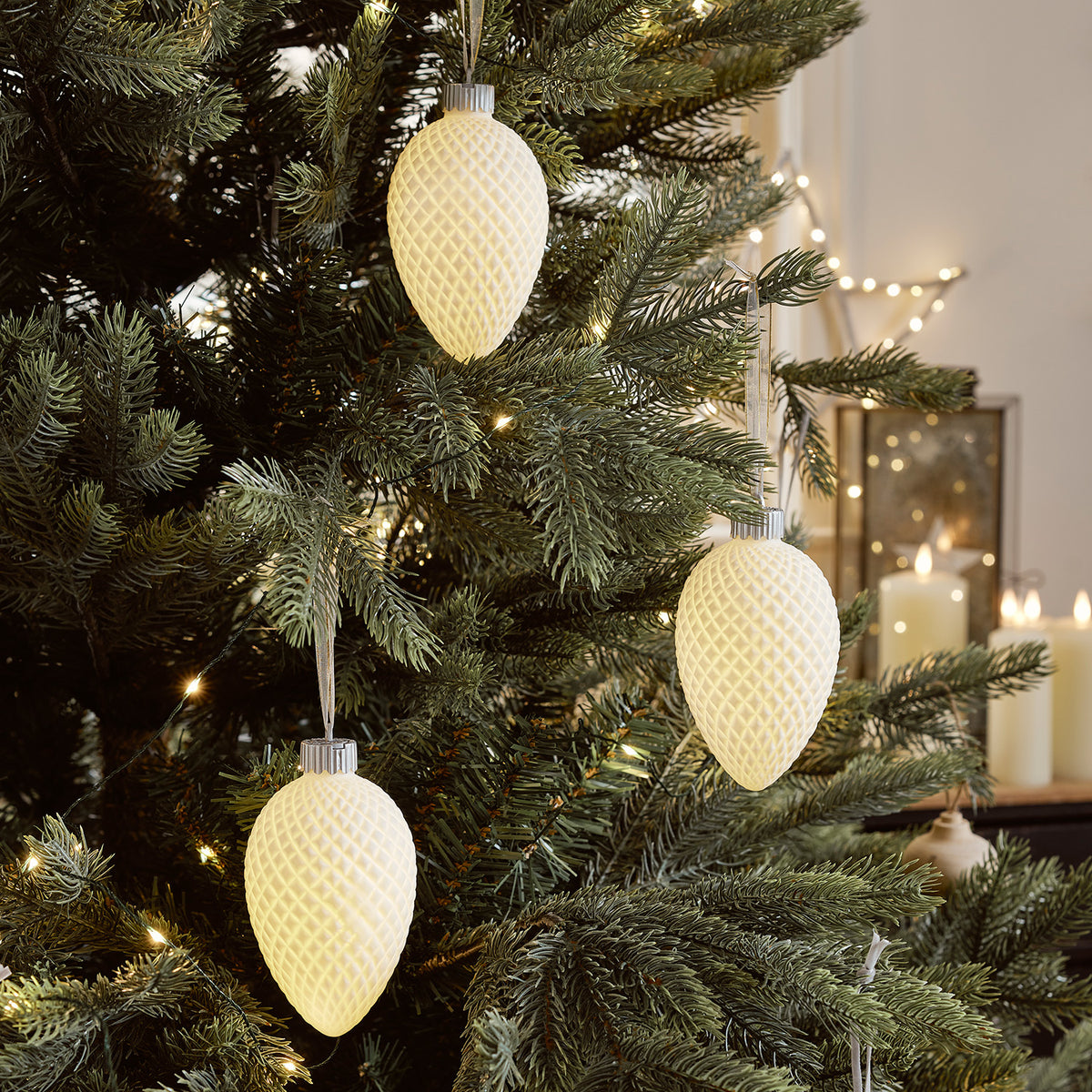 Drei leuchtende LED-Tannenzapfen aus Keramik hängen an einem Weihnachtsbaum mit Lichterkette