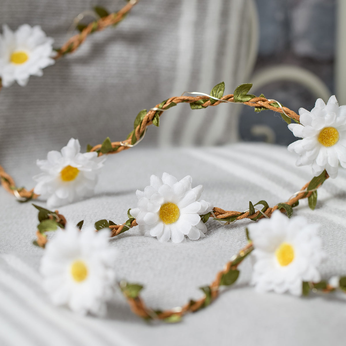 LED-Gänseblümchen-Girlande mit weißen Blüten und grünen Blättern auf hellem Stoff liegend
