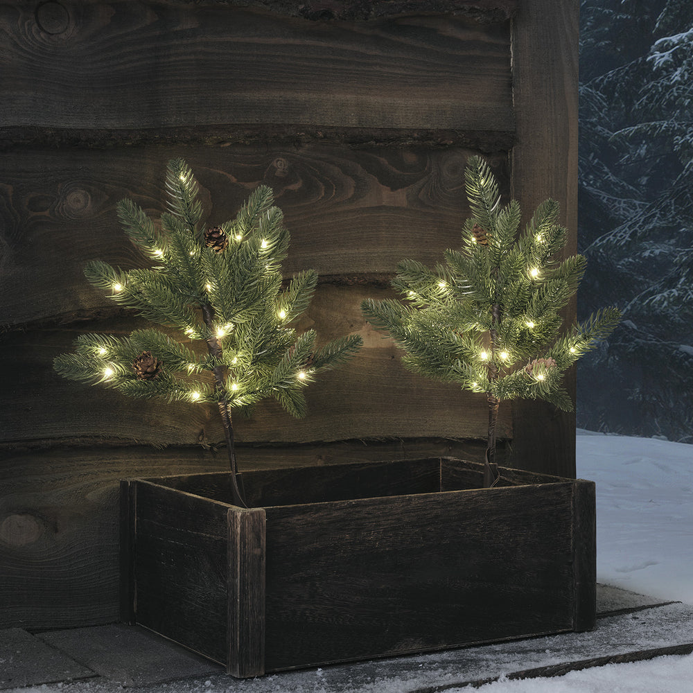 Zwei 18cm hohe LED-Tannenbäume mit Tannenzapfen und warmweißen Lichtern in einer Holzbox vor Holzwand im Schnee