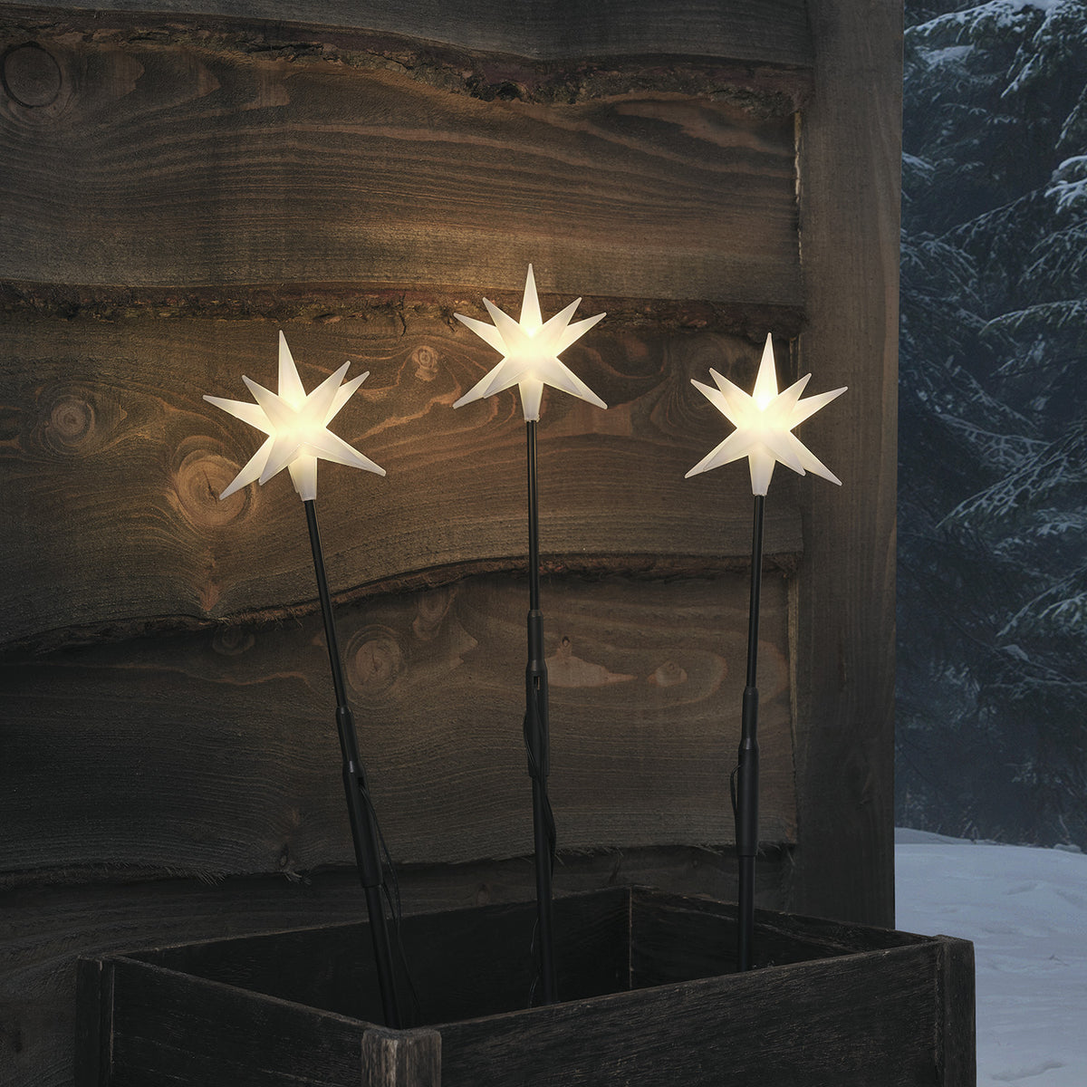 Drei beleuchtete weiße LED-Adventsterne auf schwarzen Stäben vor Holzwand und Schnee
