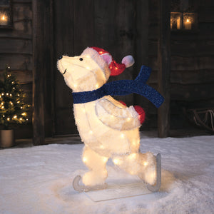 Leuchtende Weihnachtsfigur Eisbär mit roter Mütze, blauem Schal und Schlittschuhen auf Schnee vor Holzwand