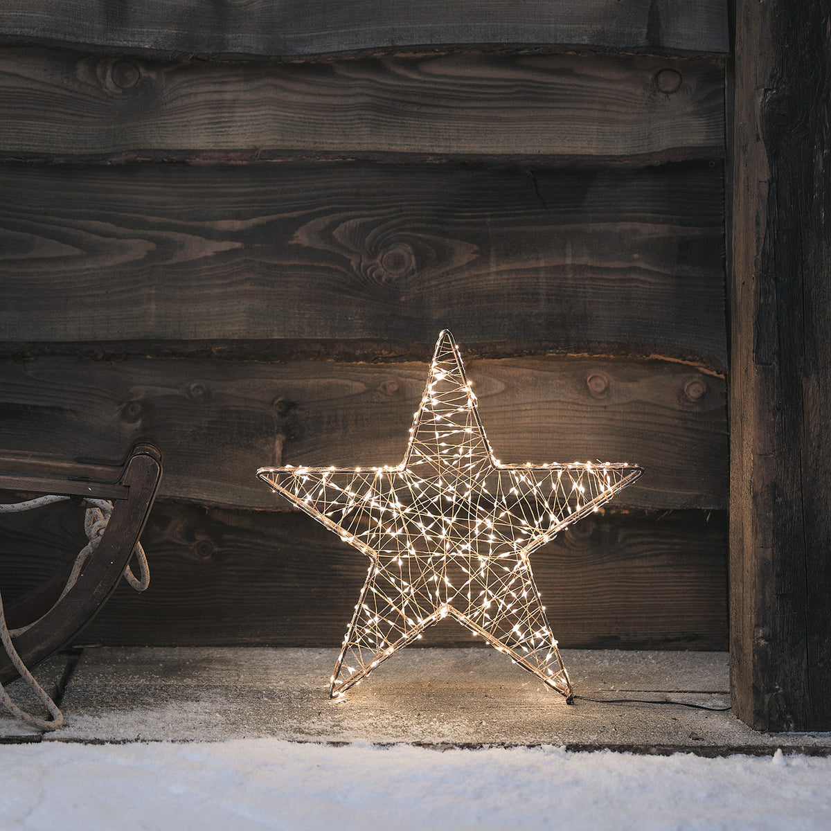 Leuchtender kupferfarbener Stern mit Micro-LEDs vor dunkler Holzwand im Schnee