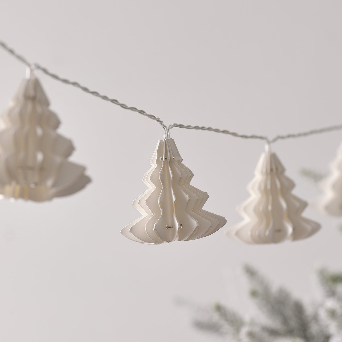 LED-Lichterkette mit weißen Papierbäumen und warmweißem Licht, unscharfer Tannenzweig im Vordergrund