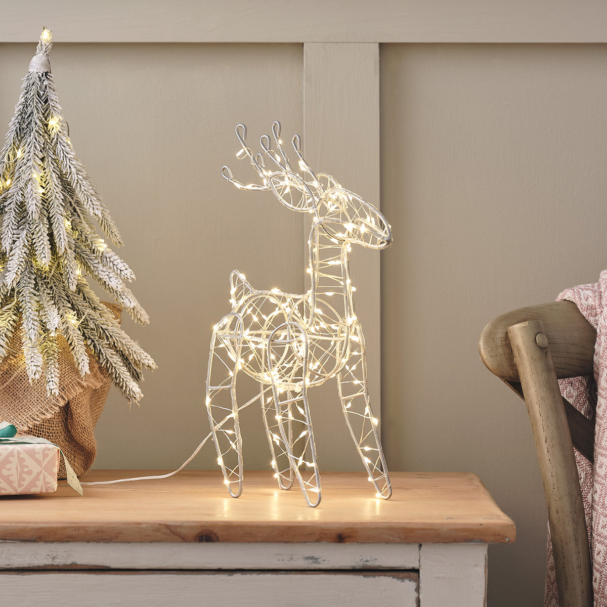 Leuchtendes Mikro-LED-Rentier aus Draht mit warmweißen LEDs auf einem Holztisch neben einem dekorierten Weihnachtsbaum