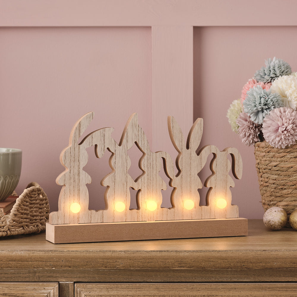 Seitenansicht der Holz-Osterhasen-Deko mit leuchtenden LEDs auf Holzsockel vor rosa Wand