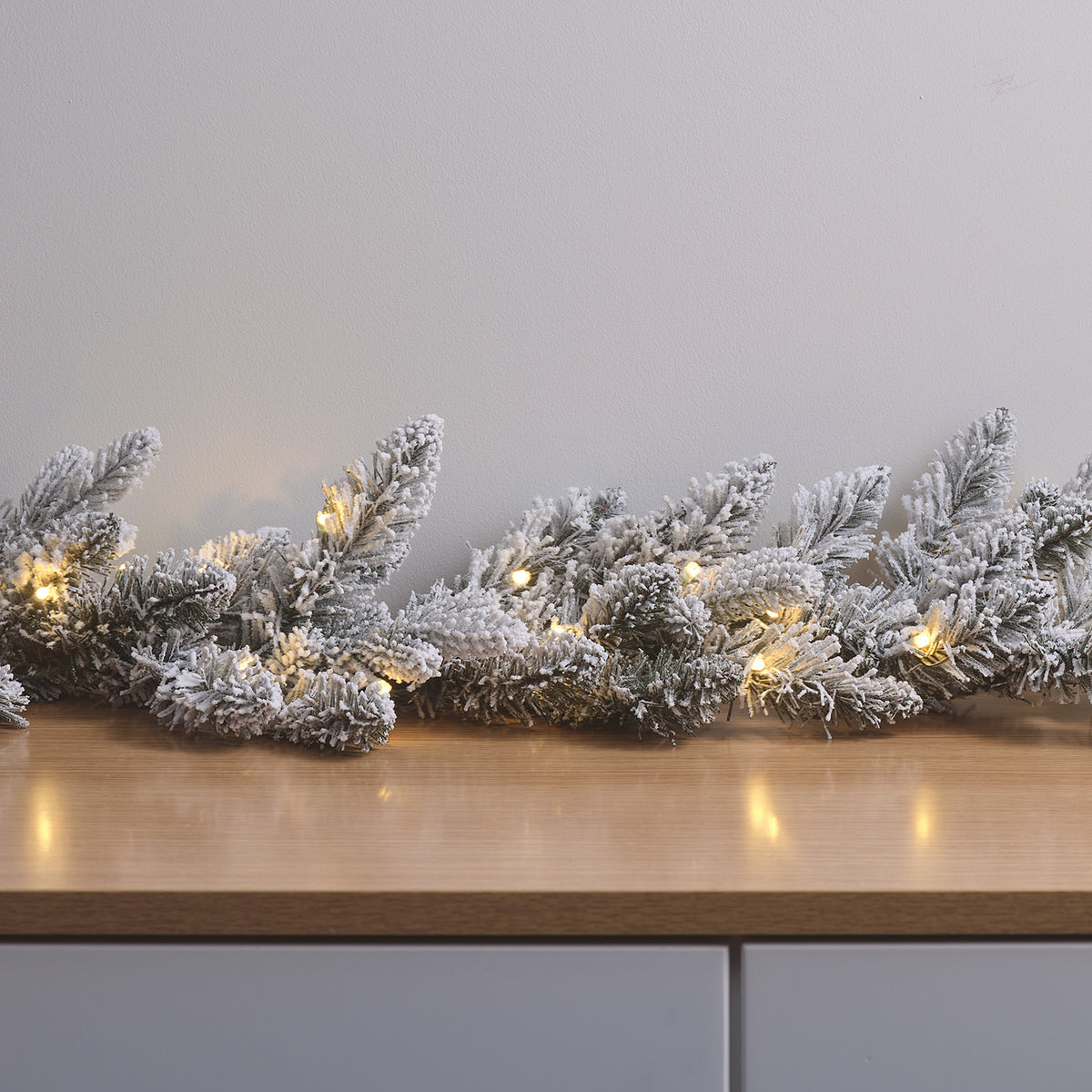 Verschneite Weihnachtsgirlande mit warmweißen LEDs auf Holzfläche vor grauer Wand