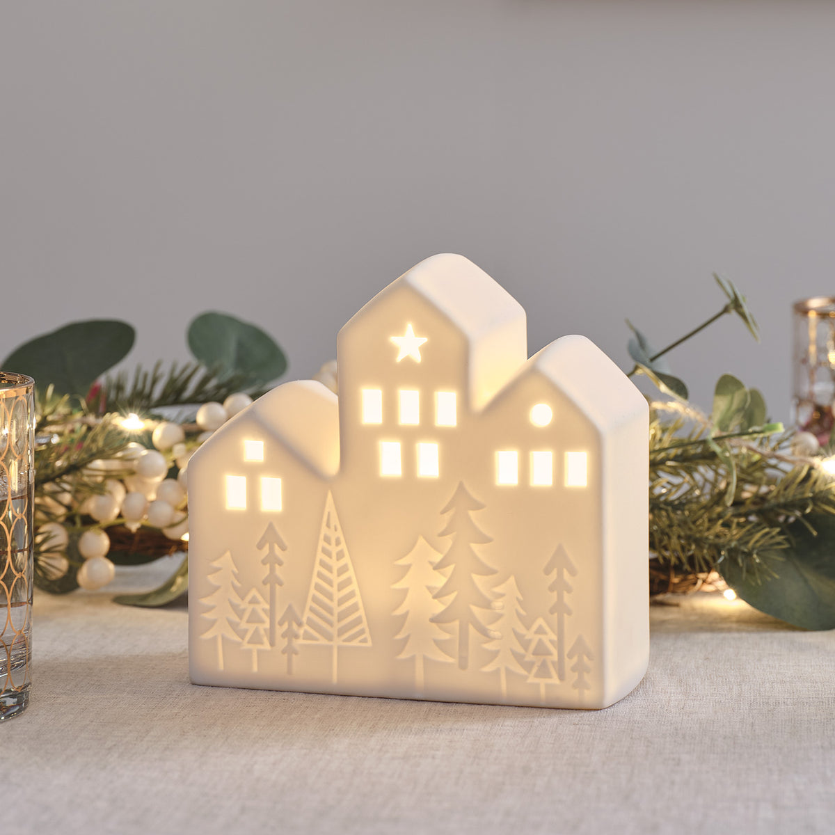 Keramikhaus mit warmweißem LED-Licht auf Tisch mit Tannenzweigen und Lichterkette