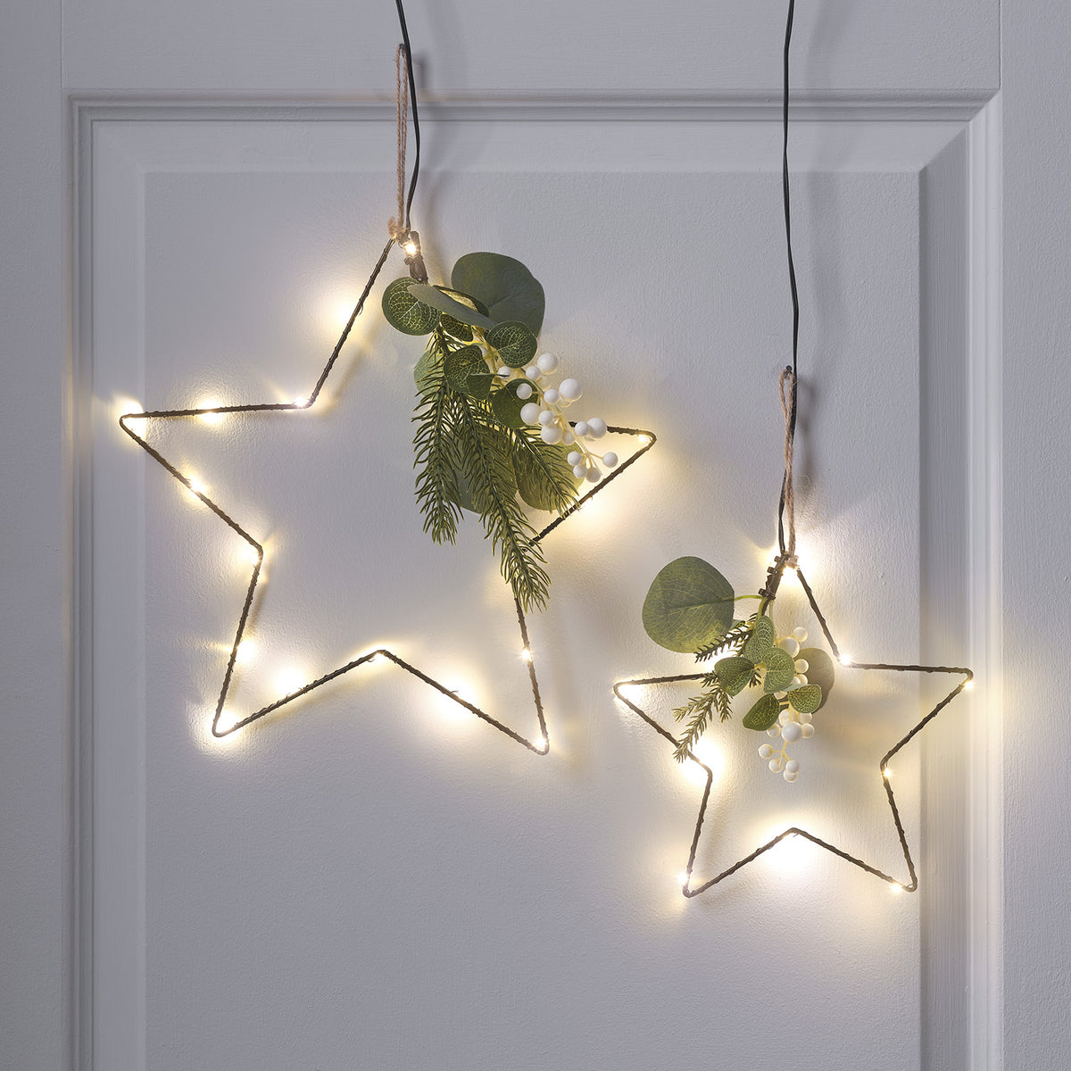 Zwei schwarze LED-Sterne mit warmweißen Lichtern und künstlichem Wintergrün hängen an einer weißen Wand