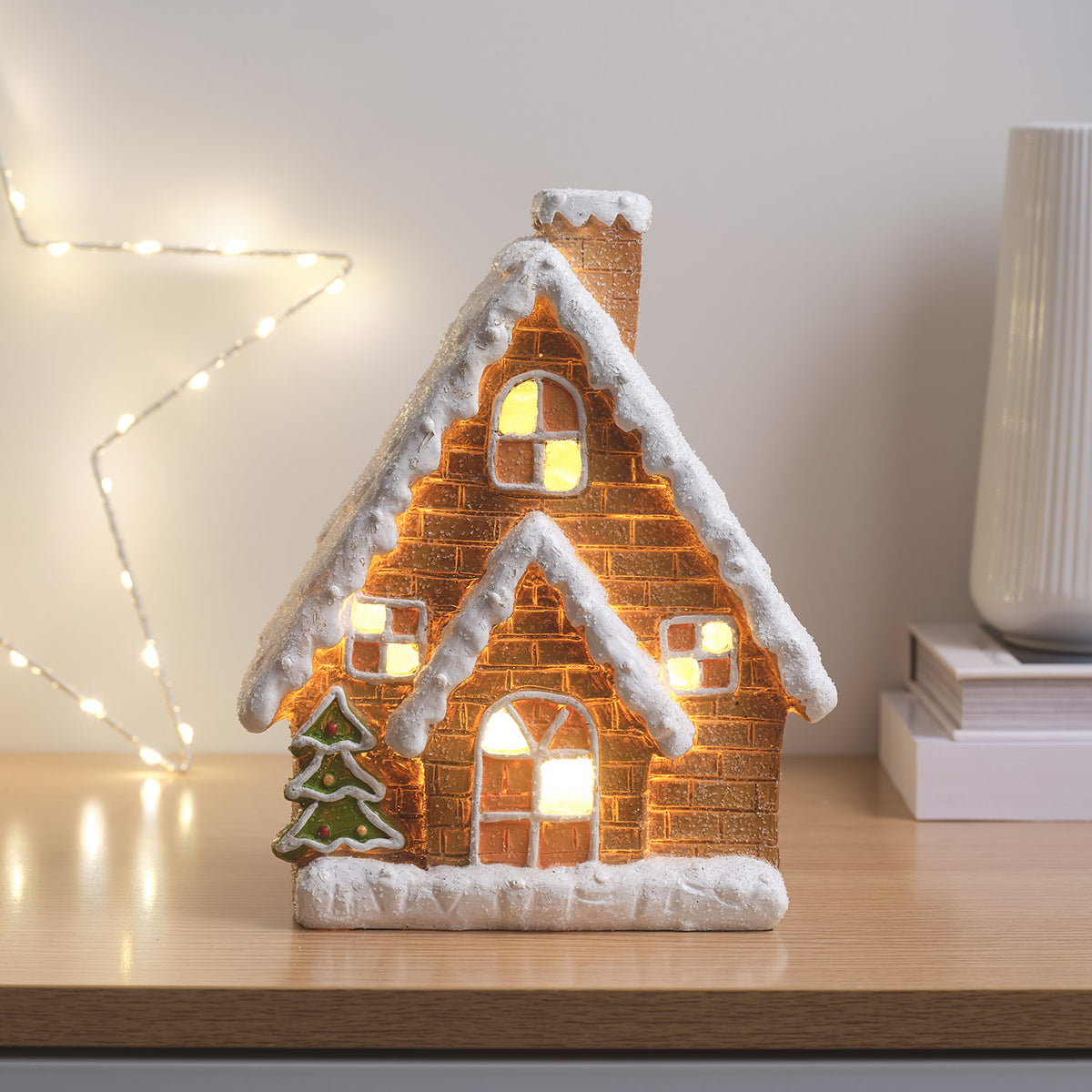 Keramik-Lebkuchenhaus mit warmweißen LEDs, Weihnachtsbaum und Schnee auf Dach, auf Holzuntergrund