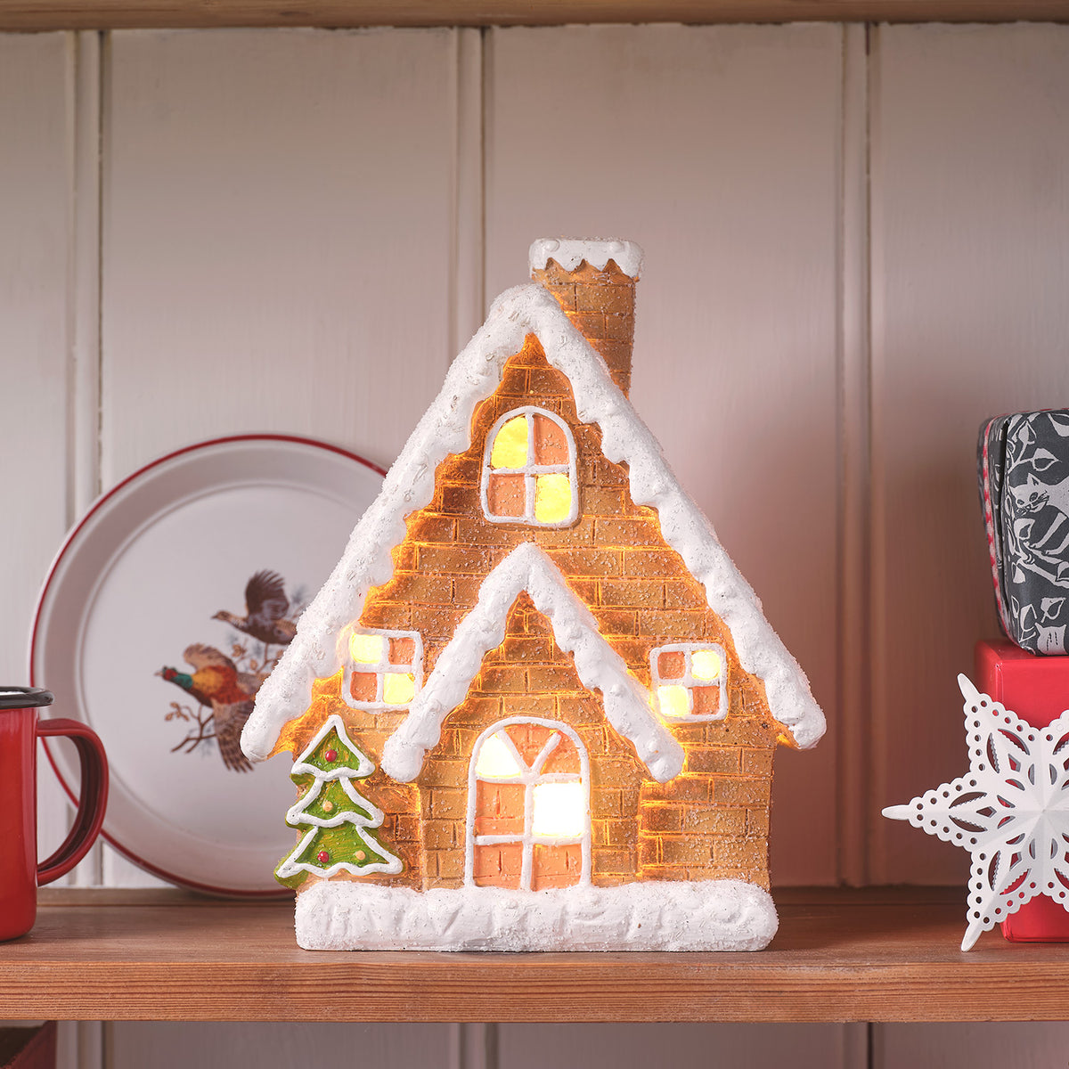 Keramik-Lebkuchenhaus mit Schnee und warmweißer LED-Beleuchtung, auf Holzplatte mit Weihnachtsdeko