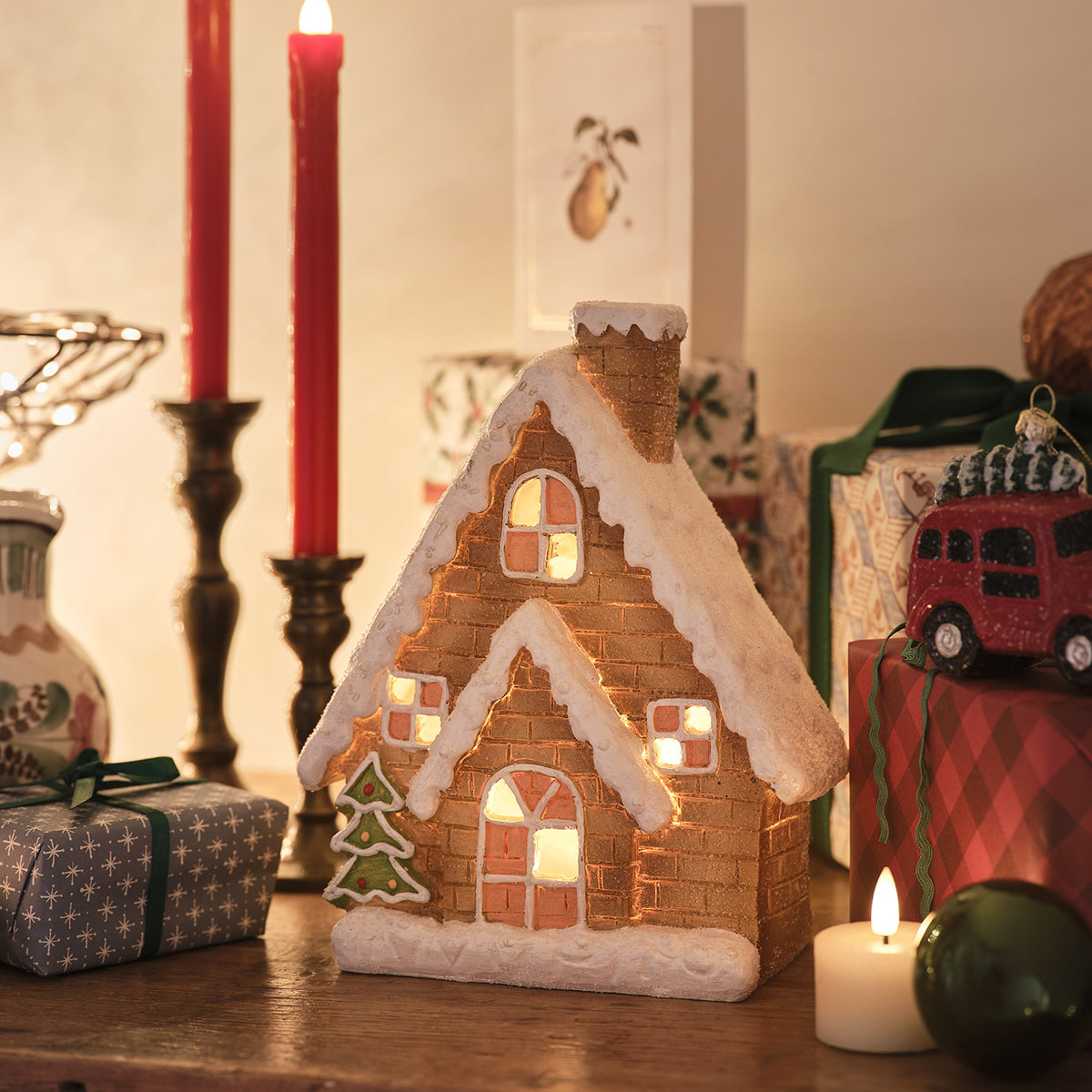 Beleuchtetes Lebkuchenhaus aus Keramik mit Weihnachtsbaum neben roten Kerzen und Geschenken