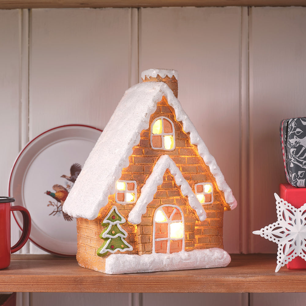 Keramik-Lebkuchenhaus mit beleuchteten Fenstern und Schnee-Dekor auf Holzregal