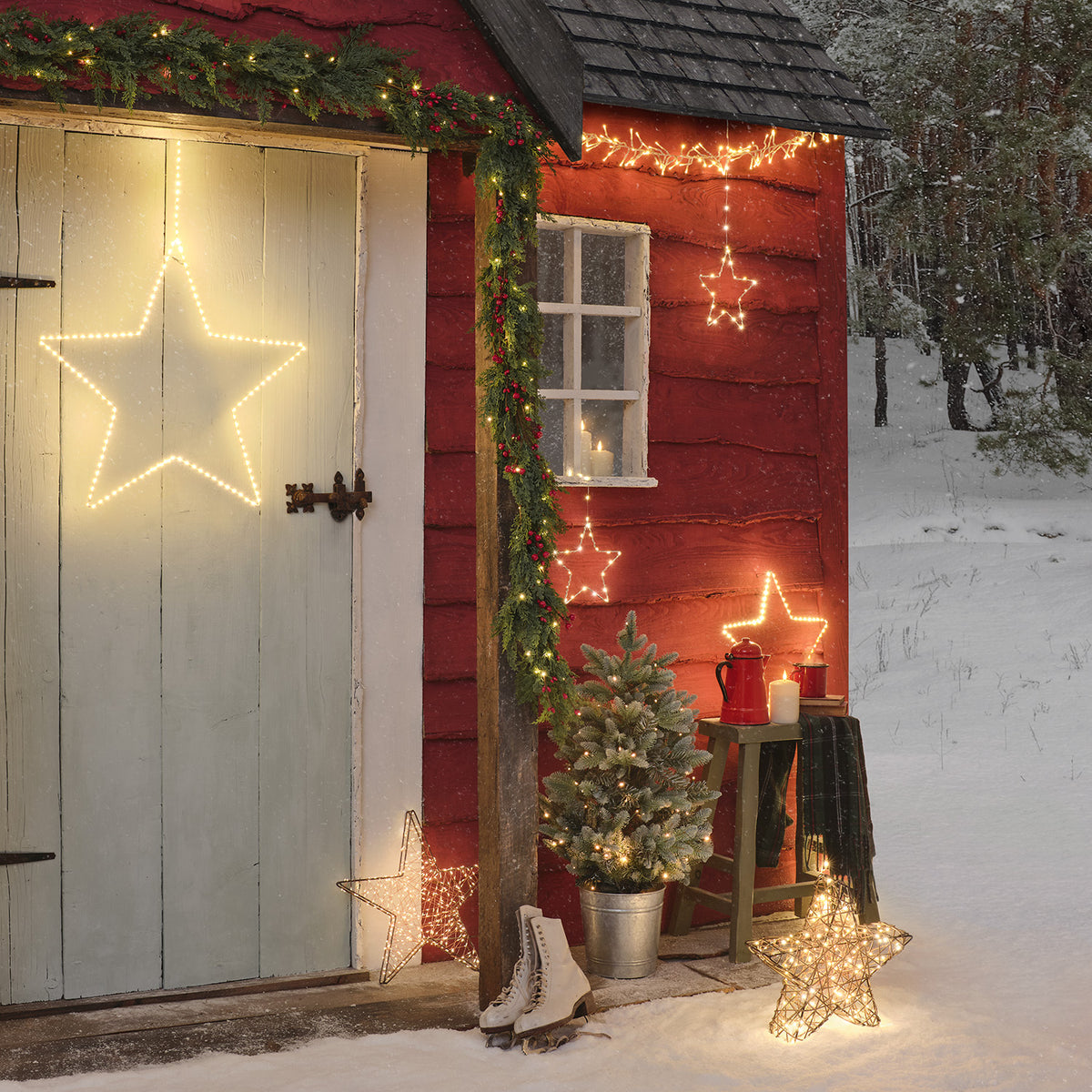 Eingang eines roten Gartenhauses mit LED-Sternen, Tannenzweigen und Schlittschuhen im Schnee