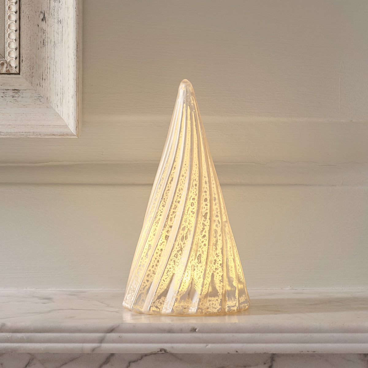 Leuchtender LED-Weihnachtsbaum aus Glas auf Marmorplatte vor weißer Wand und Bilderrahmen