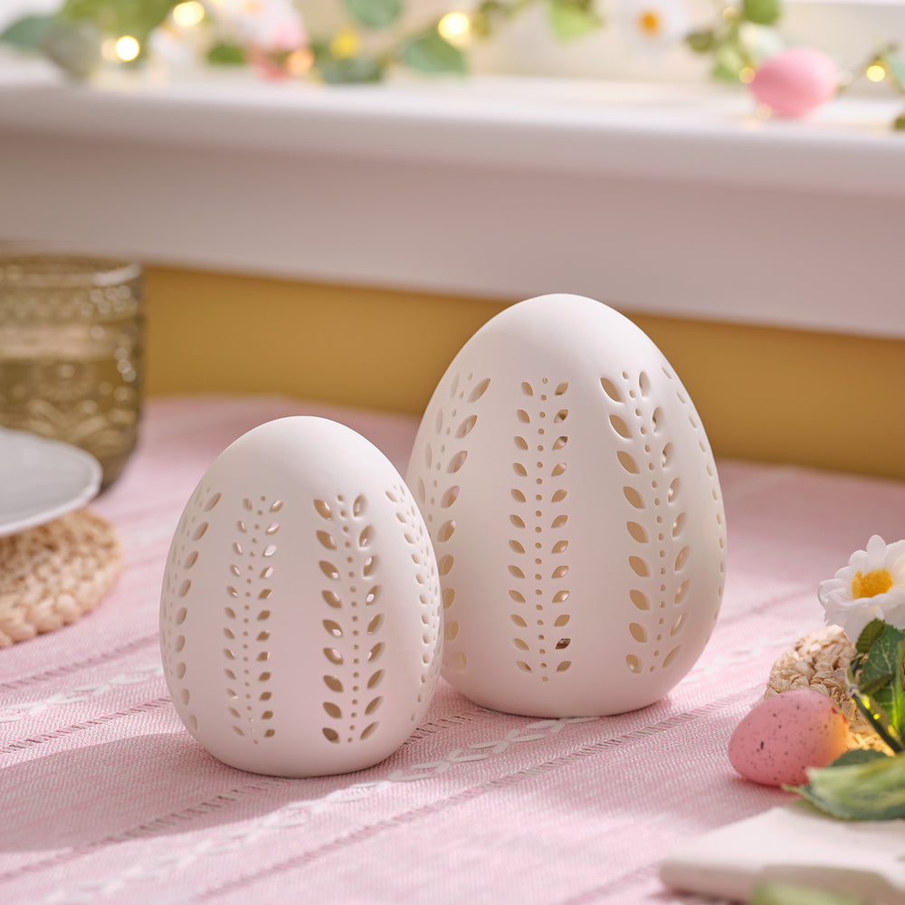 Zwei matte weiße Porzellan-Eier mit ausgestanzten Blattmustern ohne Beleuchtung auf rosa Tischdecke