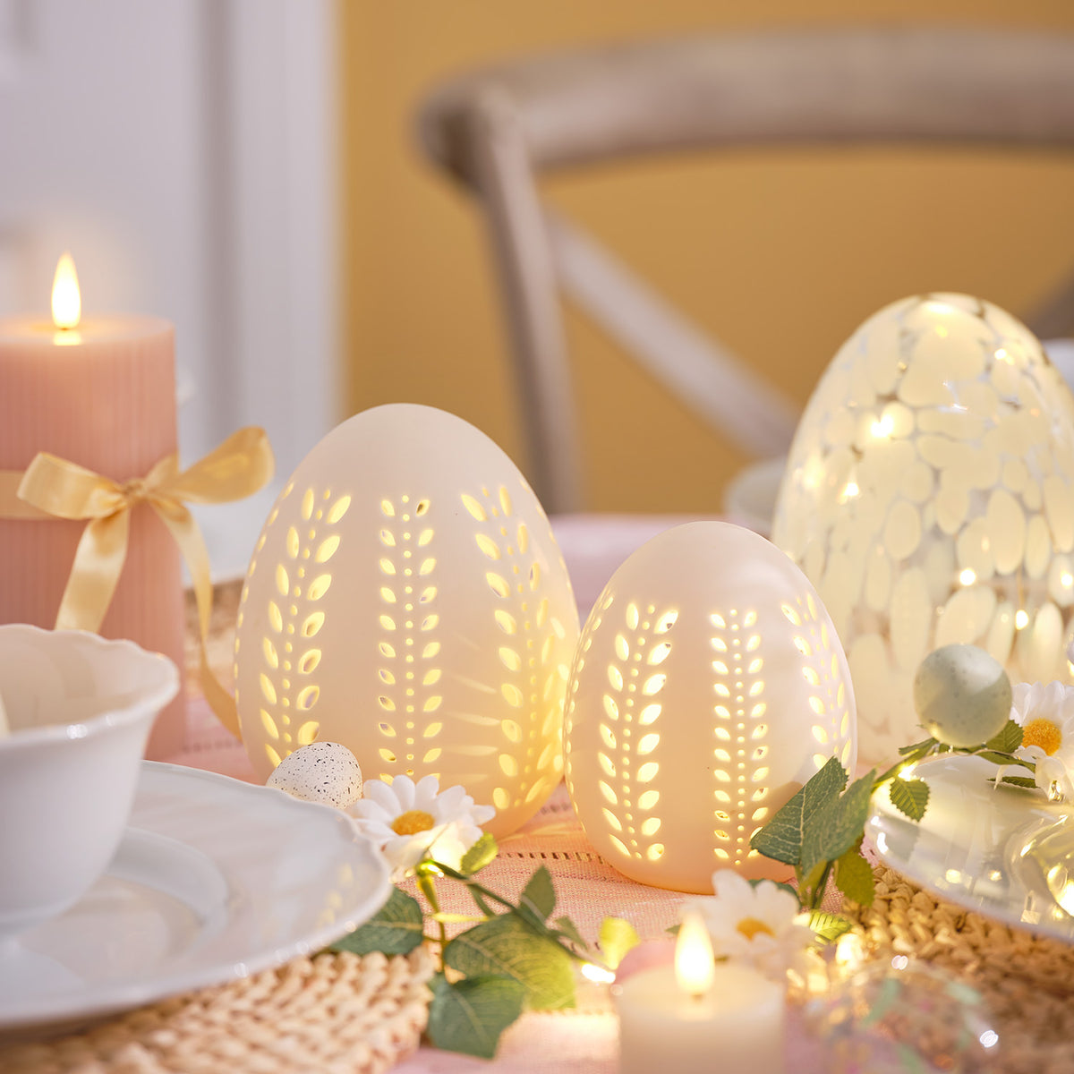 Beleuchtete LED-Porzellan-Eier mit Blattmuster auf gedecktem Ostertisch mit Kerzen und Blumen