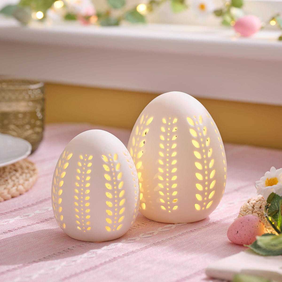 Zwei weiße Porzellan-Eier mit filigranen Blattmustern und warmweißem LED-Licht auf rosa Tischdecke