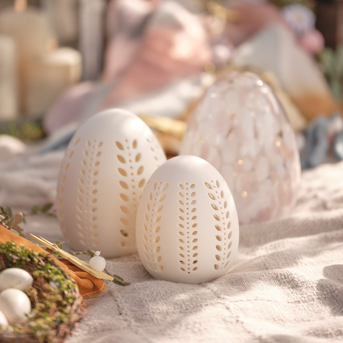 Zwei leuchtende Porzellan-Eier mit Blattmuster auf Stofftischdecke mit Osterdeko im Hintergrund