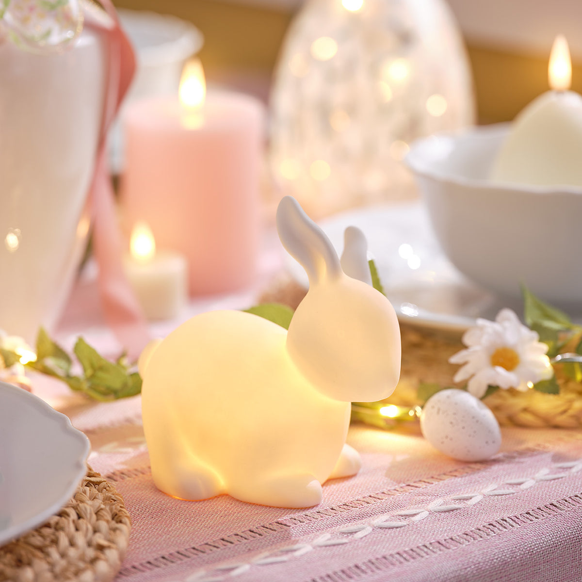 Porzellan-Osterhase mit warmweißem Licht neben Osterkerzen und Deko auf Holz