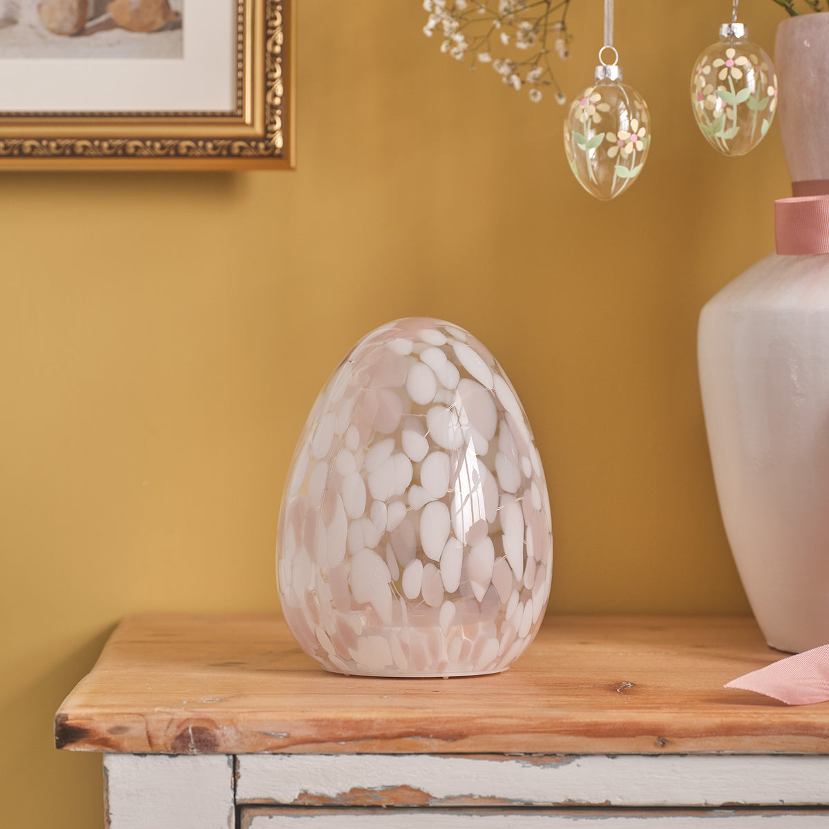 LED-Osterei aus Glas mit Konfetti-Effekt auf Holzkommode neben rosa Vase
