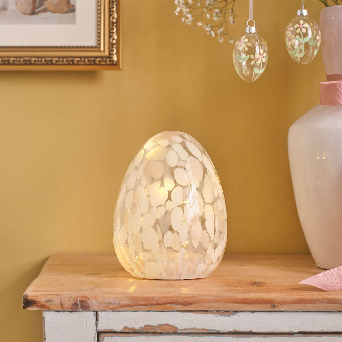 Leuchtendes Glas-Osterei mit weißen Sprenkeln auf Holzkommode vor gelber Wand