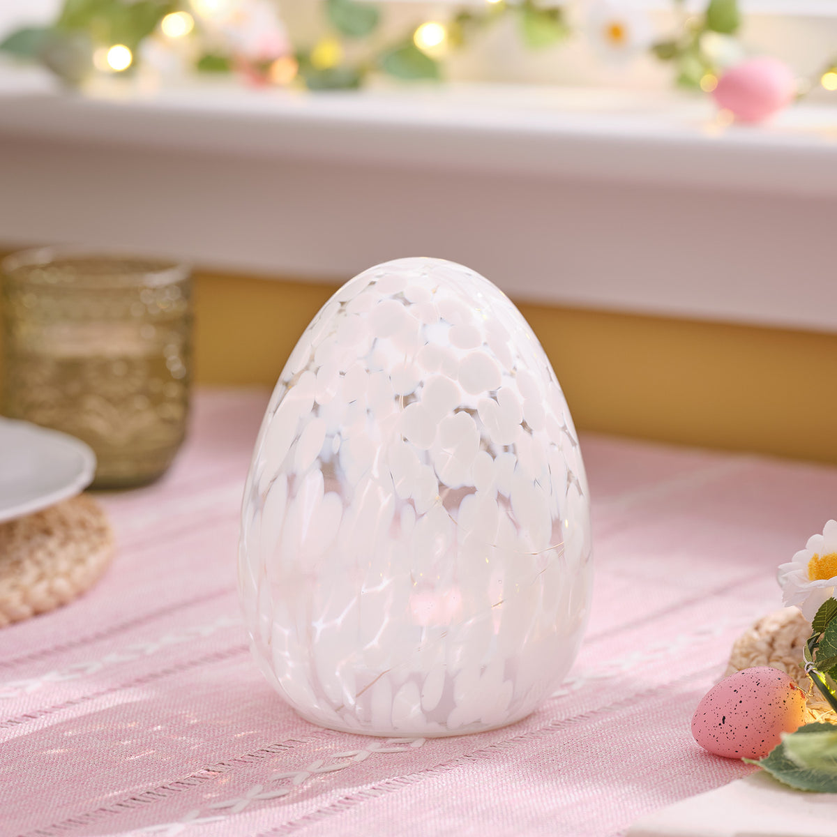 Nahaufnahme LED-Osterei aus Glas mit weißem Konfettieffekt auf rosa Tischdecke