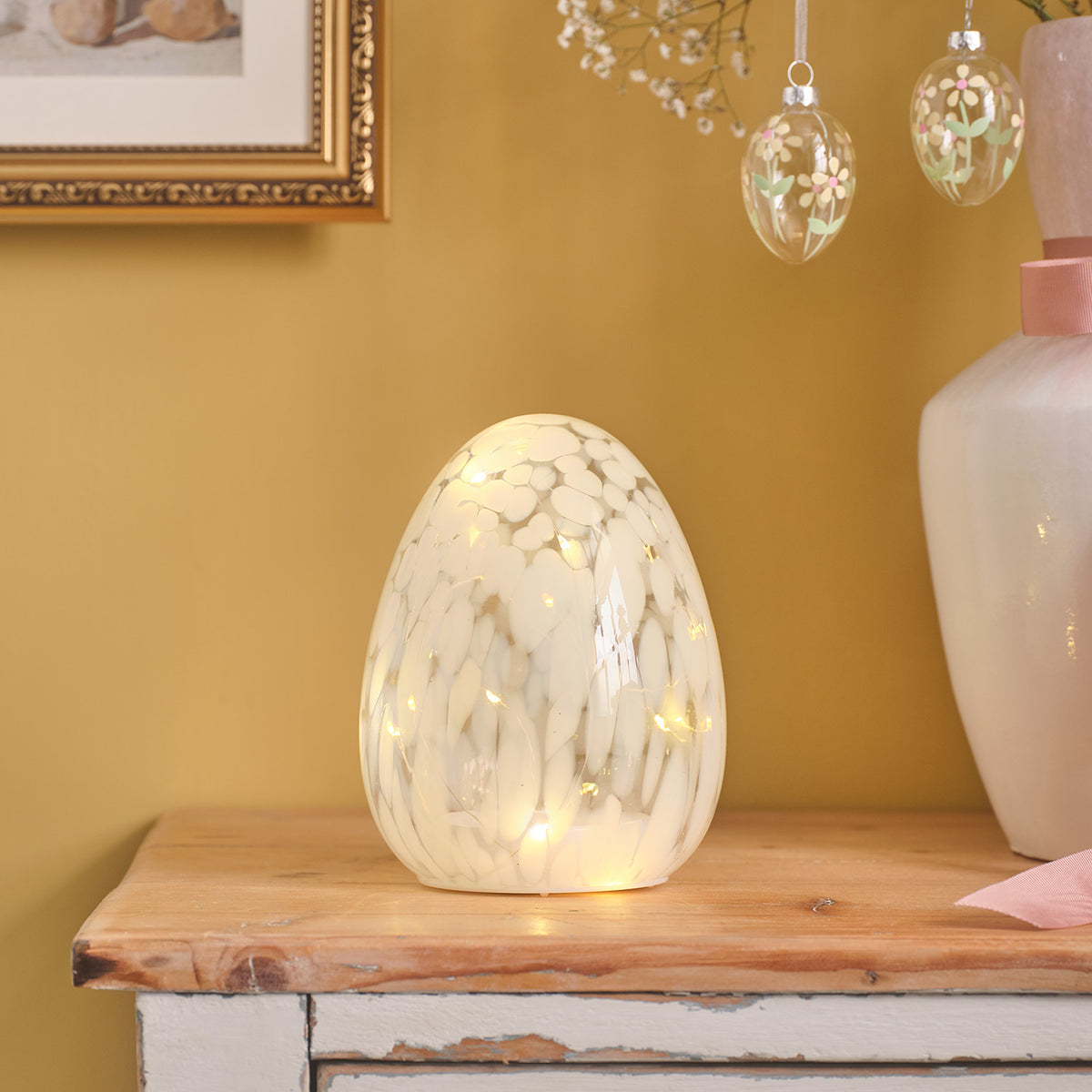Weißes LED-Osterei aus Glas mit Konfettieffekt auf Holzkommode vor gelber Wand