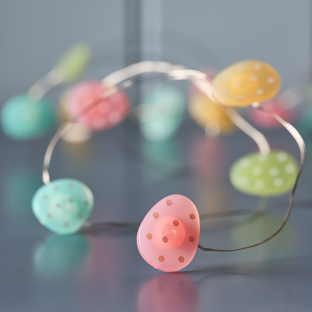 Pastellfarbene Ostereier mit weißen Punkten an transparenter LED-Lichterkette auf dunklem Untergrund