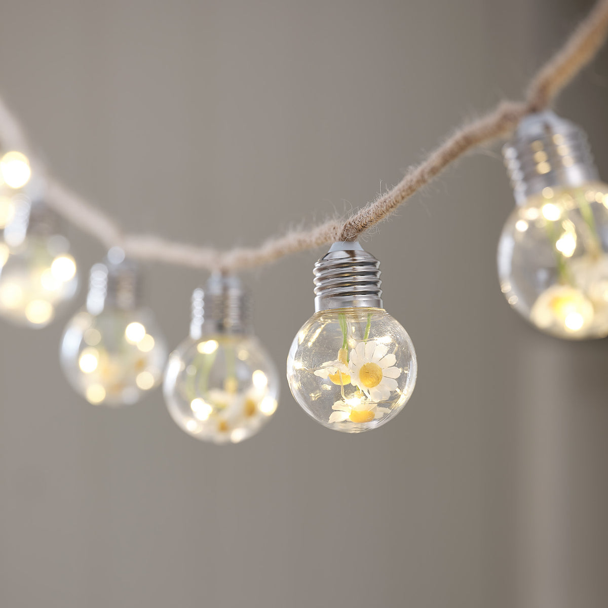 LED-Lichterkette mit Gänseblümchen in klaren Glühbirnen an rustikalem Juteseil vor hellem Hintergrund