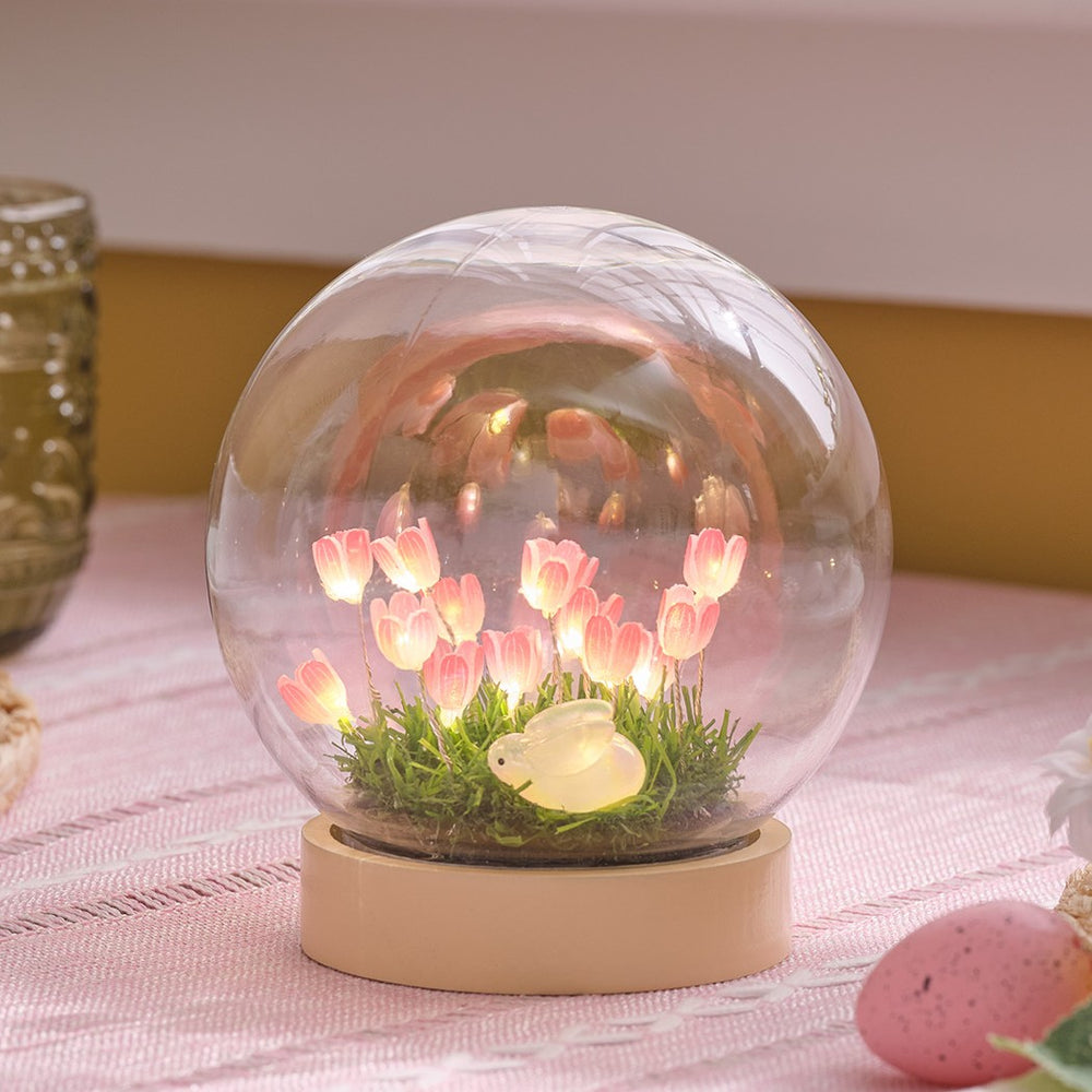 LED-Osterkugel mit beleuchteten rosa Blumen und kleinem Hasen auf grüner Wiese im Holzsockel