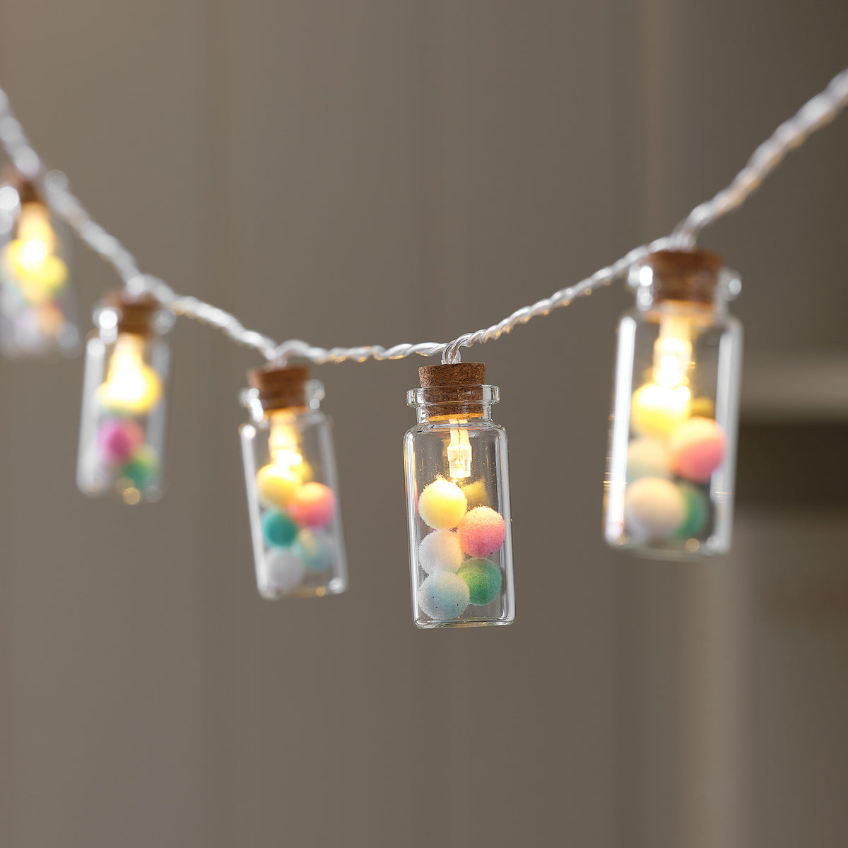 Mehrere Glasfläschchen mit bunten Pompoms und warmweiß leuchtenden LEDs an Lichterkette