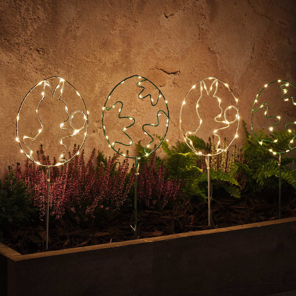 Vier LED-Osterei-Gartenstecker mit Hasen- und Blumenmotiven beleuchtet im Beet vor Wand