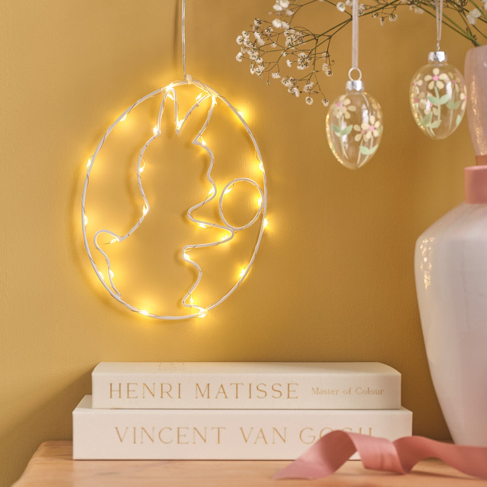 LED-Osterei mit weißem Metallrahmen und Mikro-LEDs leuchtet an gelber Wand über Büchern