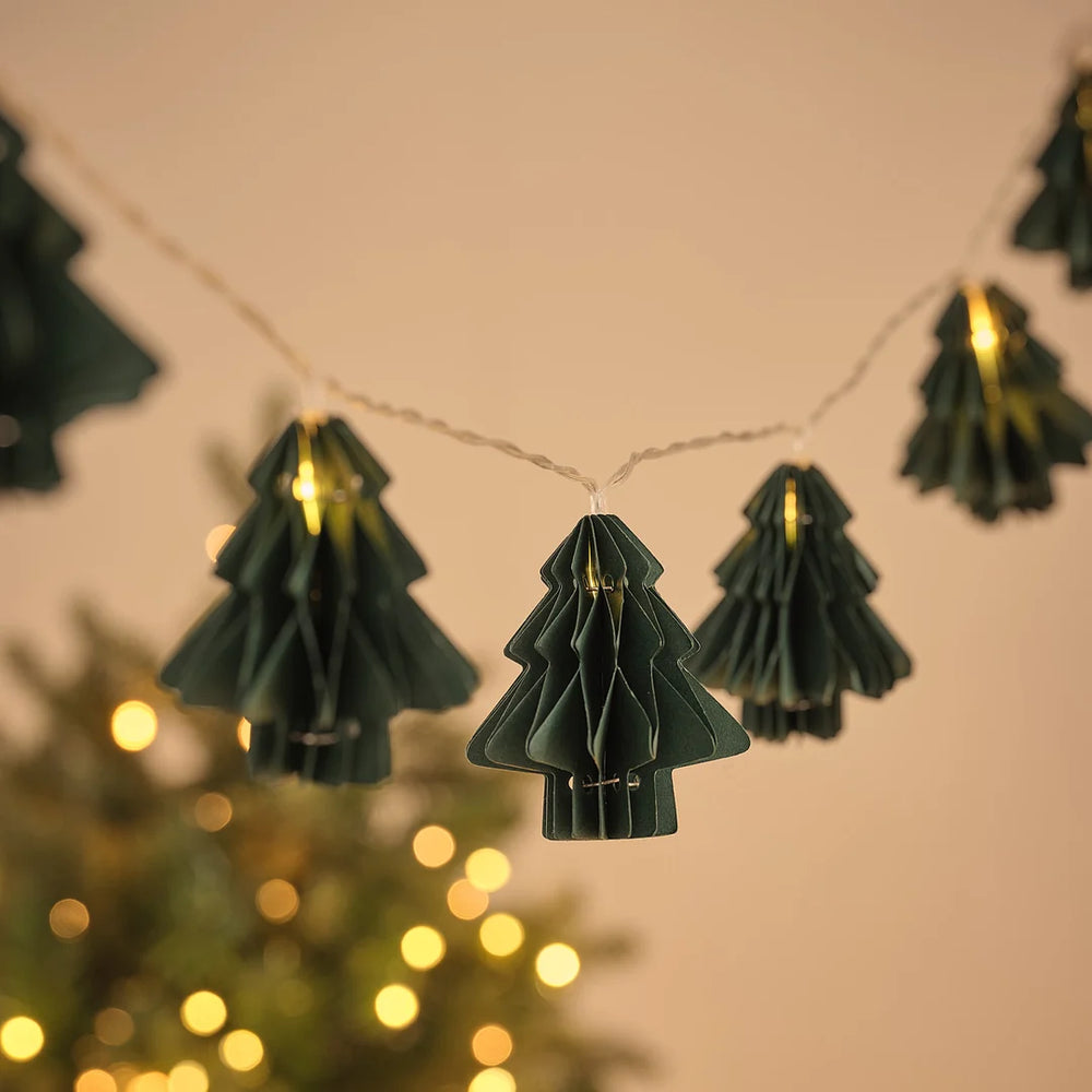 Lichterkette mit grünen Papier-Tannenbaum-Anhängern vor Weihnachtsbaum und Lichterglanz.