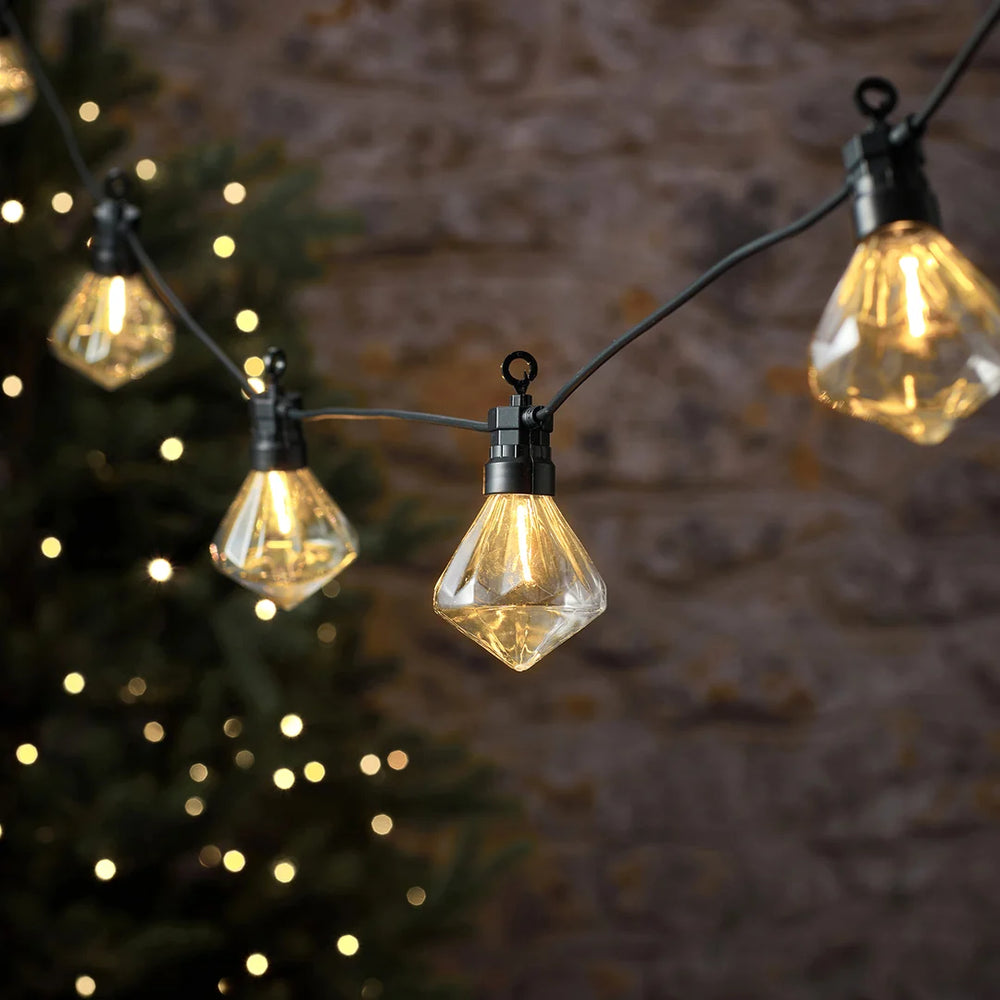 LED-Lichterkette mit diamantförmigen Glühbirnen vor Steinmauer und Tannenzweig.