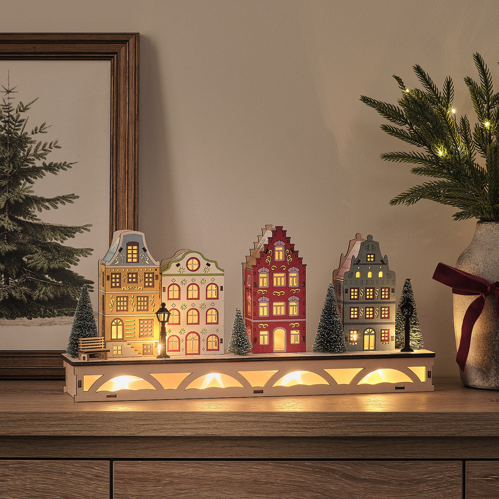 LED-Winterdorf aus Holz mit vier bunten Häuschen und schneebedeckten Bäumen beleuchtet auf Sideboard