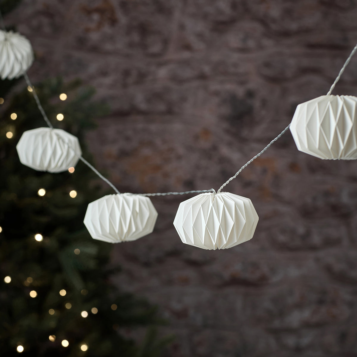 Unbeleuchtete weiße Papierlampions mit geometrischem Faltmuster an Lichterkette vor Steinwand
