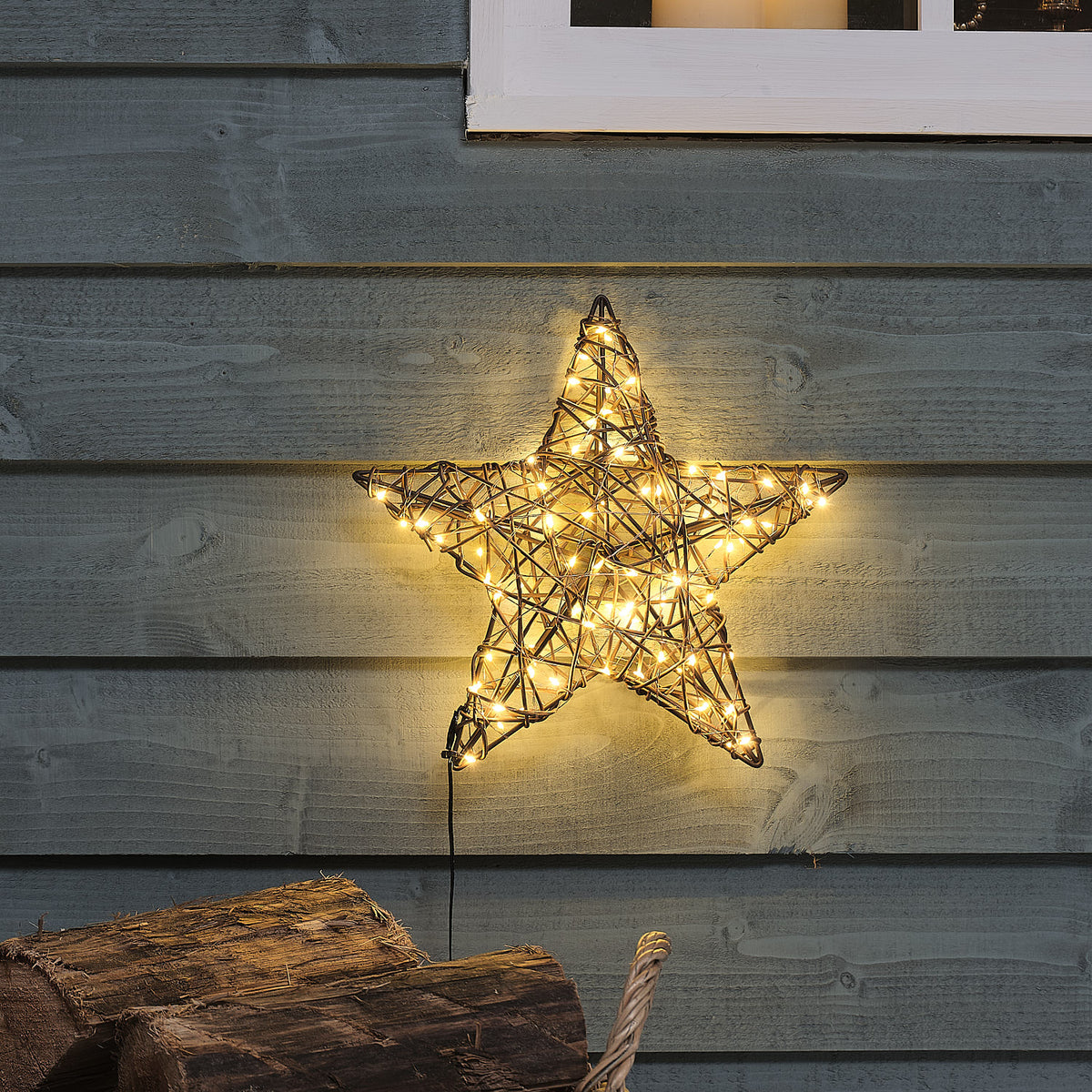 Beleuchteter Rattan-Stern mit warmweißen LEDs an blauer Holzfassade über Holzscheiten im Außenbereich
