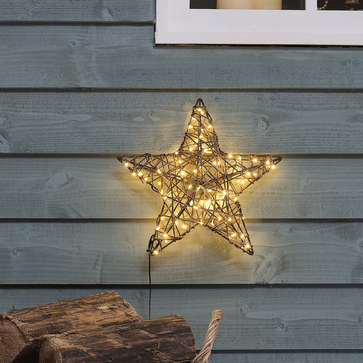 Beleuchteter Rattan-LED-Stern 30 cm an grauer Holzfassade mit Holzscheiten im Vordergrund