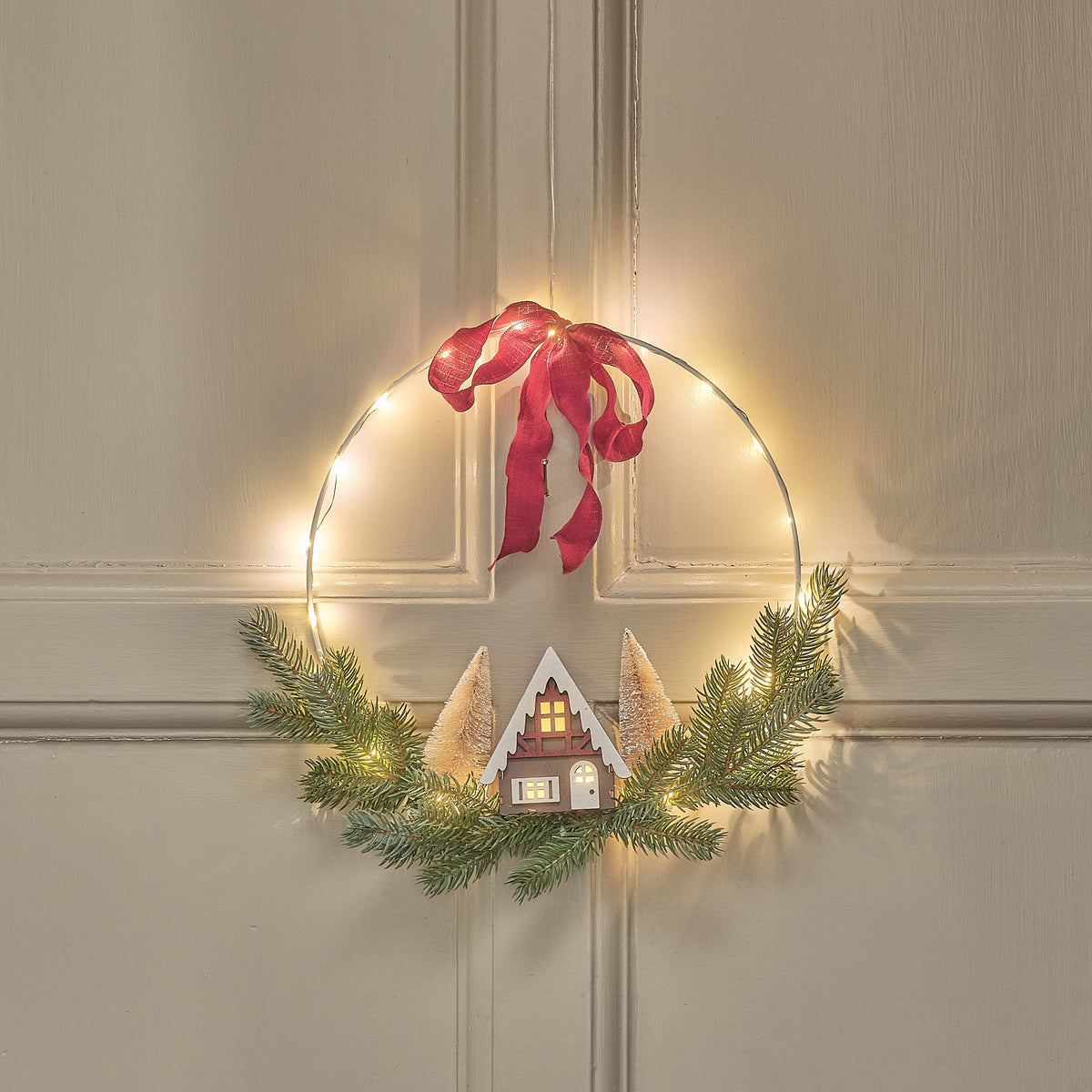 30cm LED-Weihnachtskranz mit beleuchtetem Drahtring, Häuserszene, Tannengrün und bordeauxroter Schleife an Wand