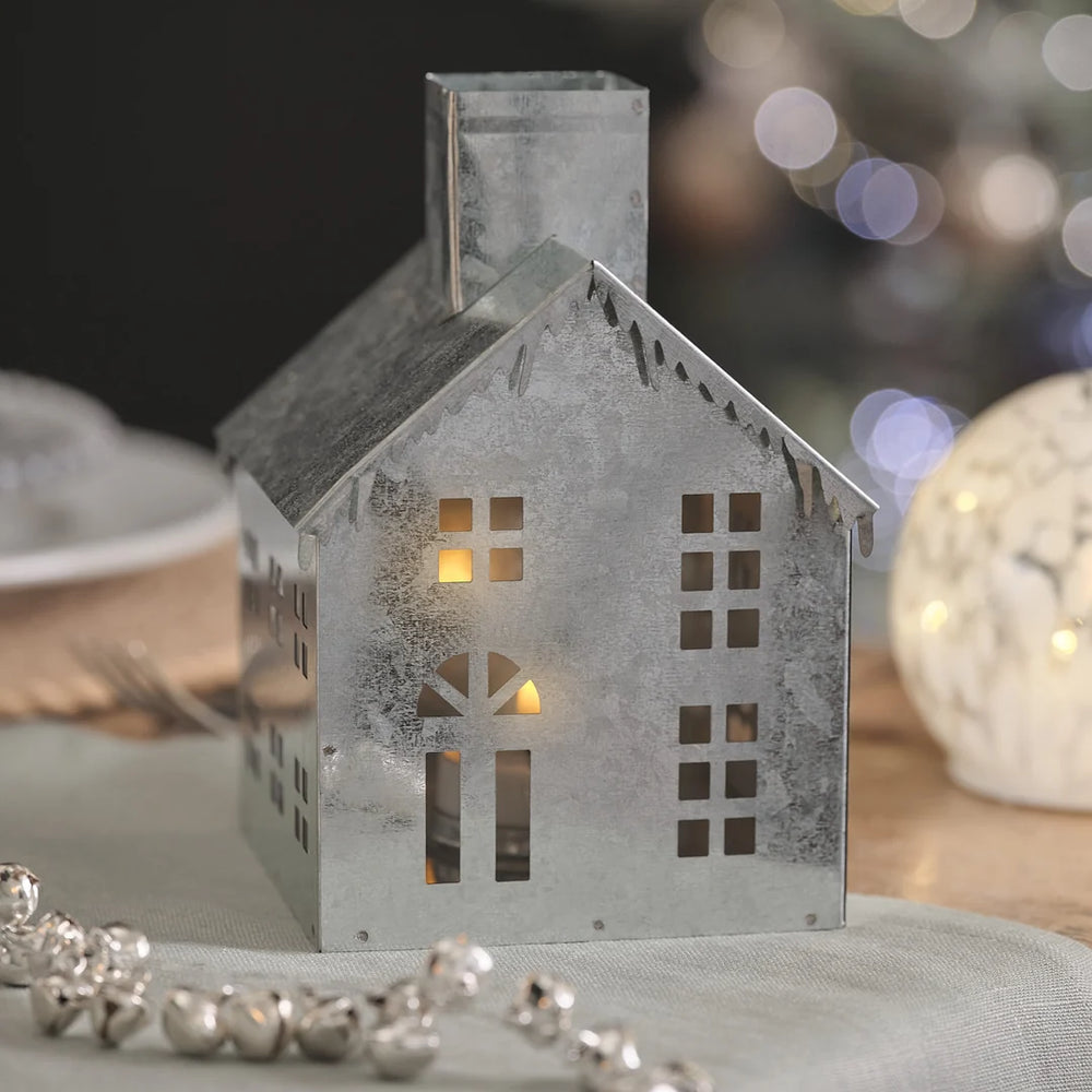 Elegantes beleuchtetes Weihnachtshaus aus grauem Porzellan mit warmweißen LED-Lichtern in den Fenstern, dekoriert mit cremefarbener Perlen-Girlande auf dunklem bokeh-Hintergrund mit unscharfen Lichtern – hochwertige LED-Dekofigur für festliche Weihnachtsarrangements, Kaminsims-Dekorationen und stimmungsvolle Raumszenen.
