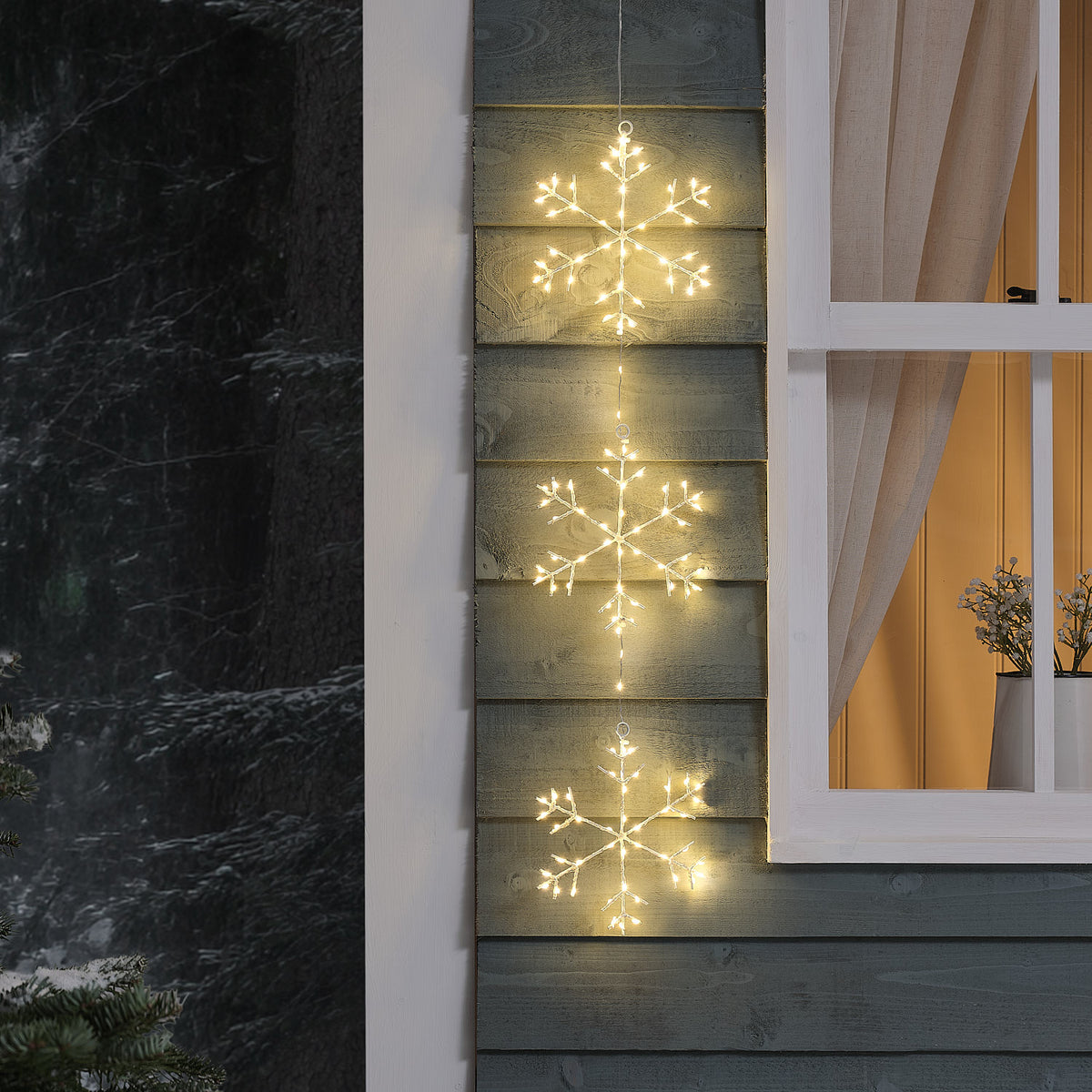 Drei hängende LED-Schneeflocken in Warmweiß an einer Hauswand neben einem Fenster