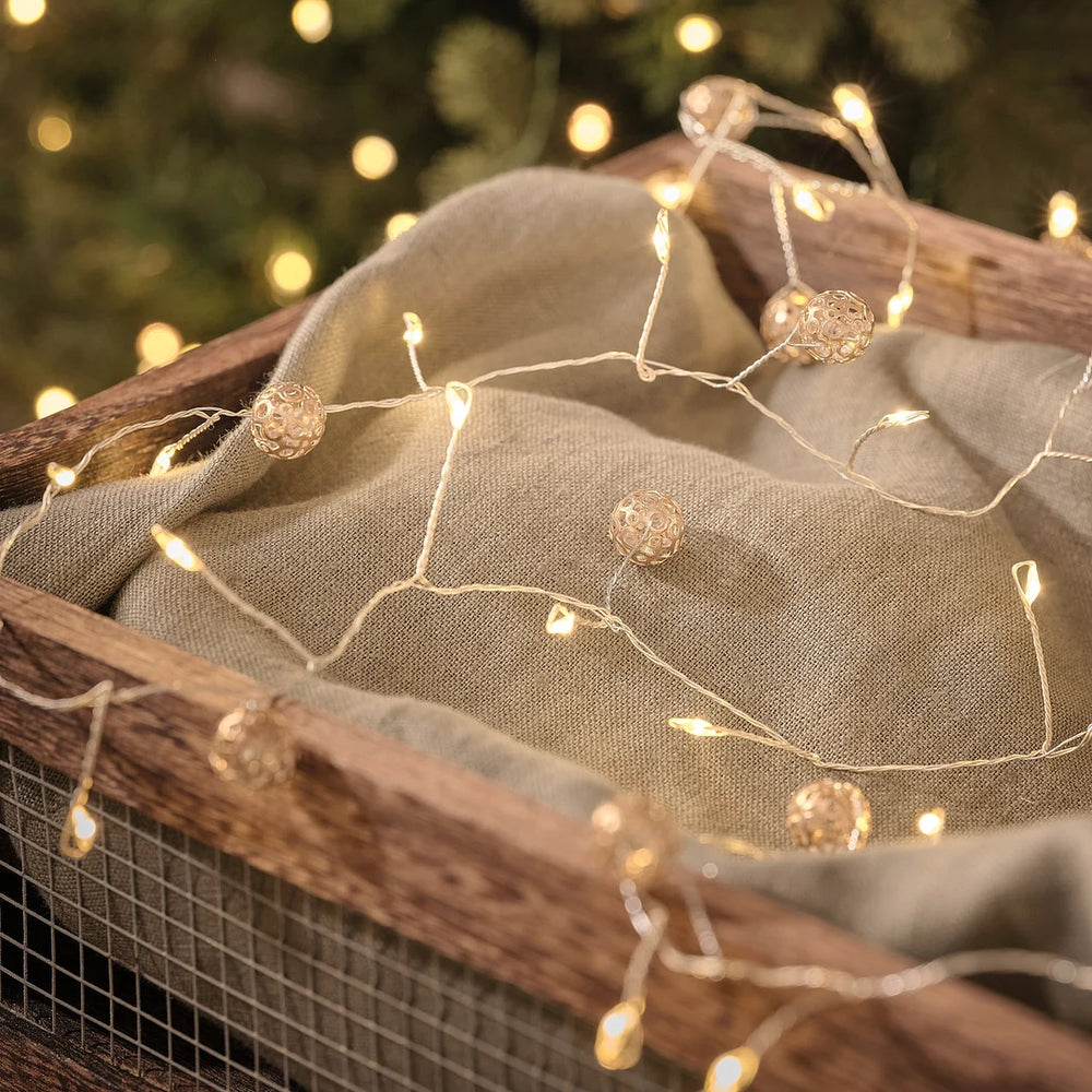 Elegante LED-Lichterkette mit warmweißen Leuchtperlen, dekoriert mit cremefarbenen Organza-Schleifen und braunen Trockenzweigen, auf neutralem Textilhintergrund – festliche Lichterkette für Weihnachtsdekoration, Fensterdekorationen und elegante Indoor-Arrangements mit gemütlichem warmweißem Glanz.