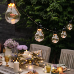 Warmweiße Party Lichterkette mit Glühbirnen über gedecktem Gartentisch bei Dämmerung