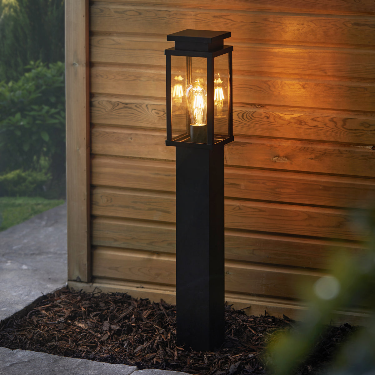 Schwarze Aluminium-Wegeleuchte mit Glaswänden leuchtet vor einer Holzverkleidung im Außenbereich